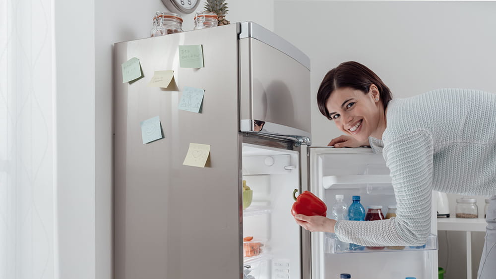 Woman taking a fresh healthy pepper from the fridge and smiling at camera