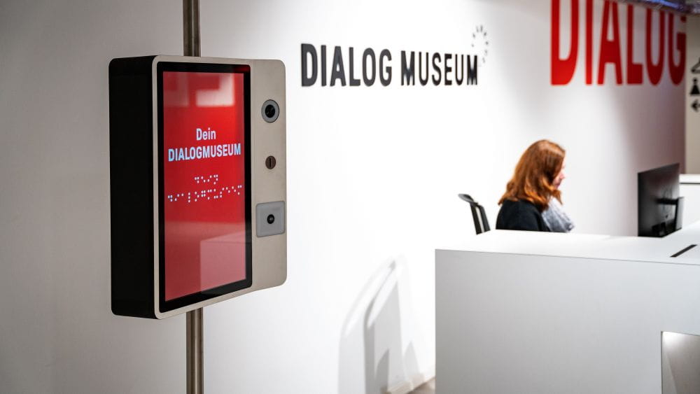 Foyer des Dialogmuseums mit Display im Vordergrund und Frau hinter dem Empfangstresen sowie dem Schriftzug des Museums