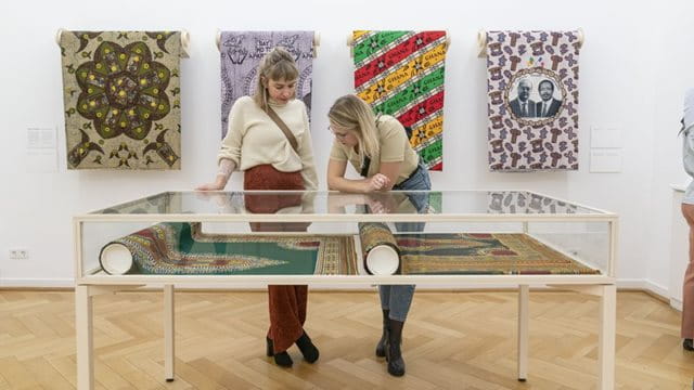 Zwei junge Frauen schauen sich eine Vitrine mit Teppichen im Weltkulturen Museum an, hinter ihnen an der Wand hängen Wandteppiche