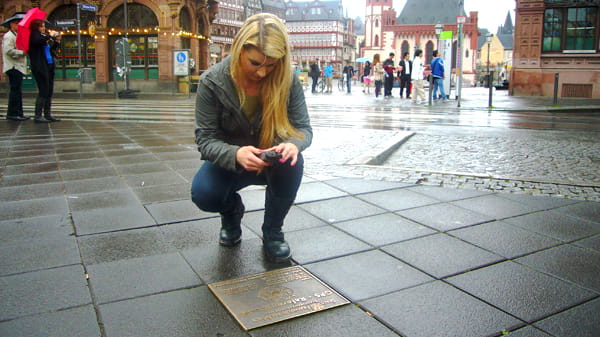 Eine junge, blonde Frau kniet vor der Bodenplatte, die am GPS-Referenzpunkt in den Boden eingelassen ist. Sie blickt auf ihren GPS Empfänger. 