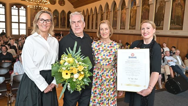 Die Verleihung des Binding-Kulturpreises im Kaisersaal des Frankfurter Römers Vier Personen stehen im Kaisersaal des Römers, dahinter sitzt das Publikum. Die Frau auf der rechten Seite des Bildes hält die Auszeichnung Binding-Kulturpreis 2025 in der Hand.