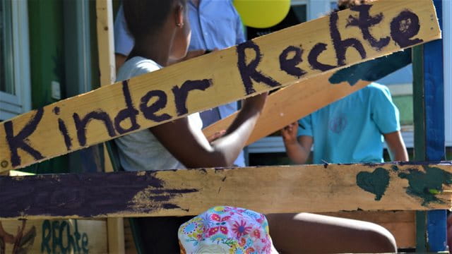 Eine Holzplanke wurde mit den Worten Kinder und Rechte bemalt. Im Hintergrund sind Kinder zu sehen.