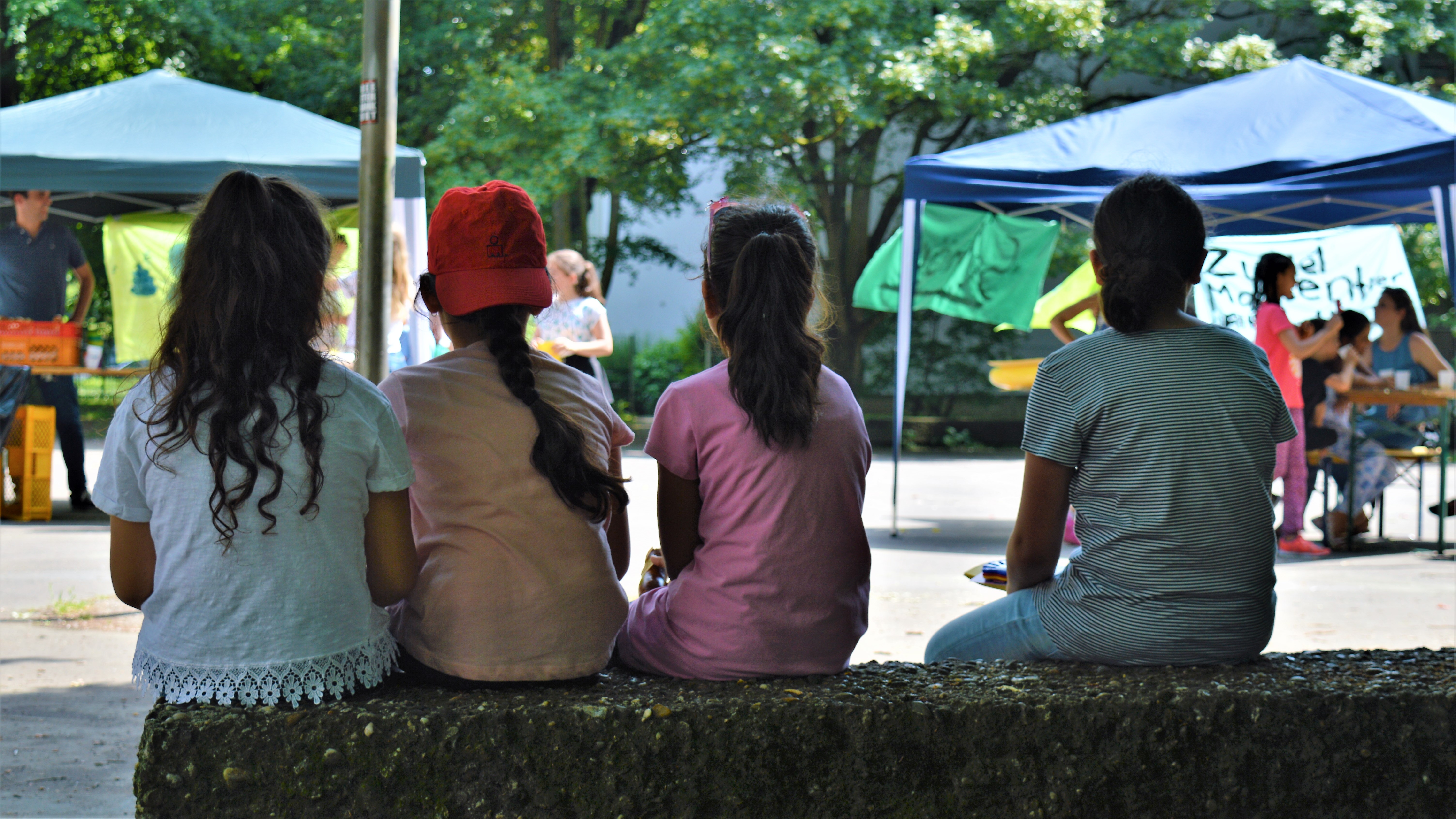 Vier Mädchen sitzen im Schatten einer Platane und beobachten eine Veranstaltung.