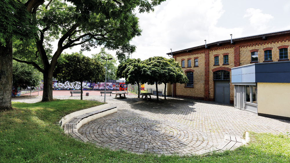 Blick über das Außengelände des Jugend Kulturzentrums mit viel Grün, Möglichkeiten für Tischtennis, Fußball, Basketball und Sitzgelegenheiten