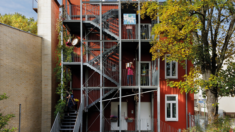 Blick auf das Kinderhaus Griesheim von außen, die Mitarbeiter sind auf den einzelnen Stockwerken der Treppe mit unterschiedlichen Posen zu sehen. 