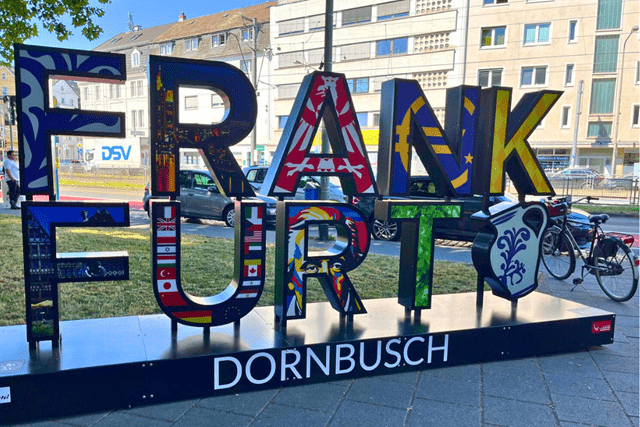 Frankfurt Selfie-Point im Stadtteil Dornbusch Große bunte Buchstaben vor blauem Himmel, grünem Baum und einer vielbefahrenen Straße.