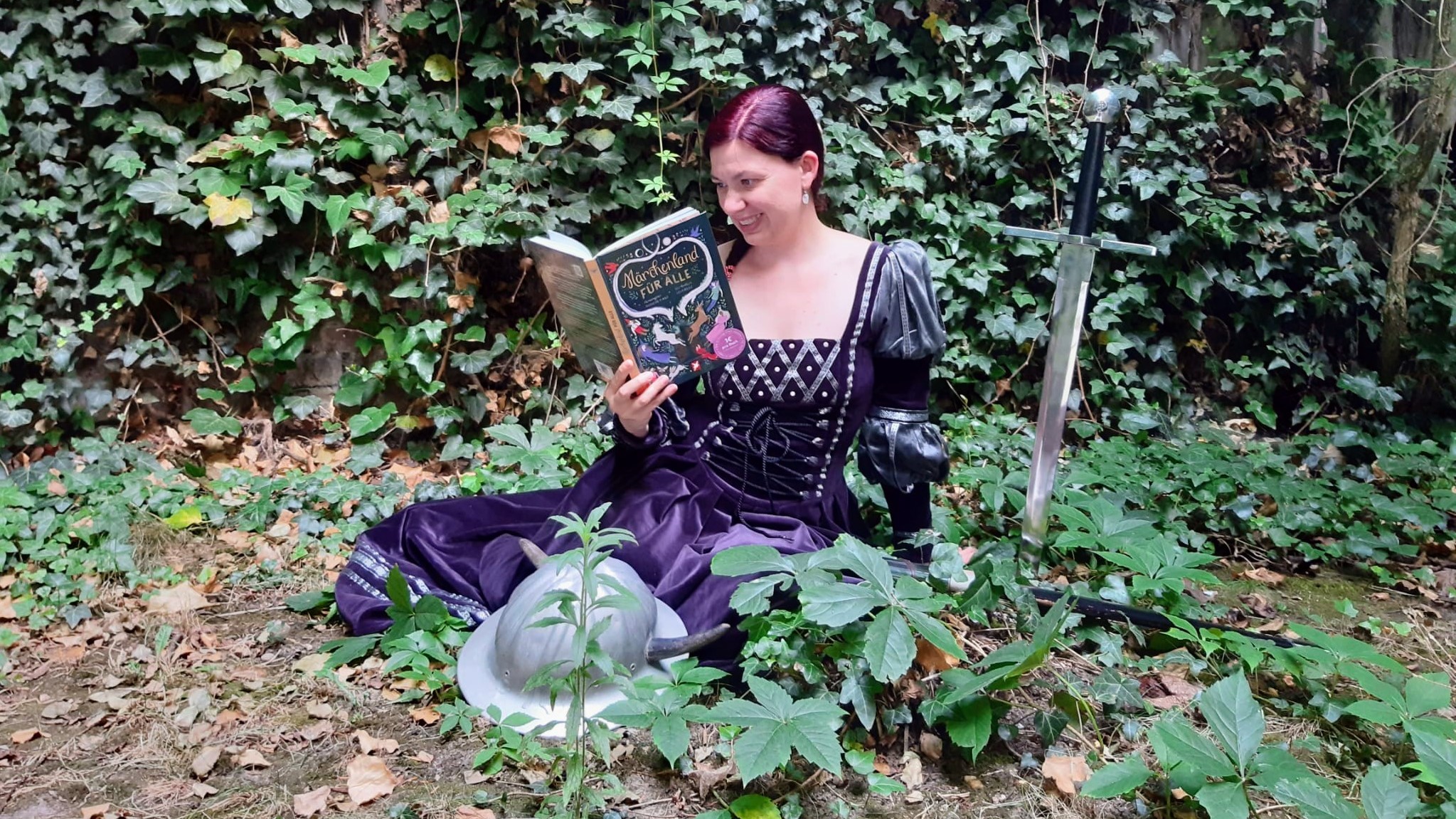 Frau in Märchenkleid sitzt im Wald neben einem Schwert und liest "Märchenland für Alle".