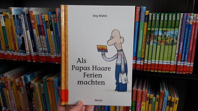 Das Buch "Als Papas Haare Ferien machten" vor einem Regal mit anderen Kinderbüchern.
