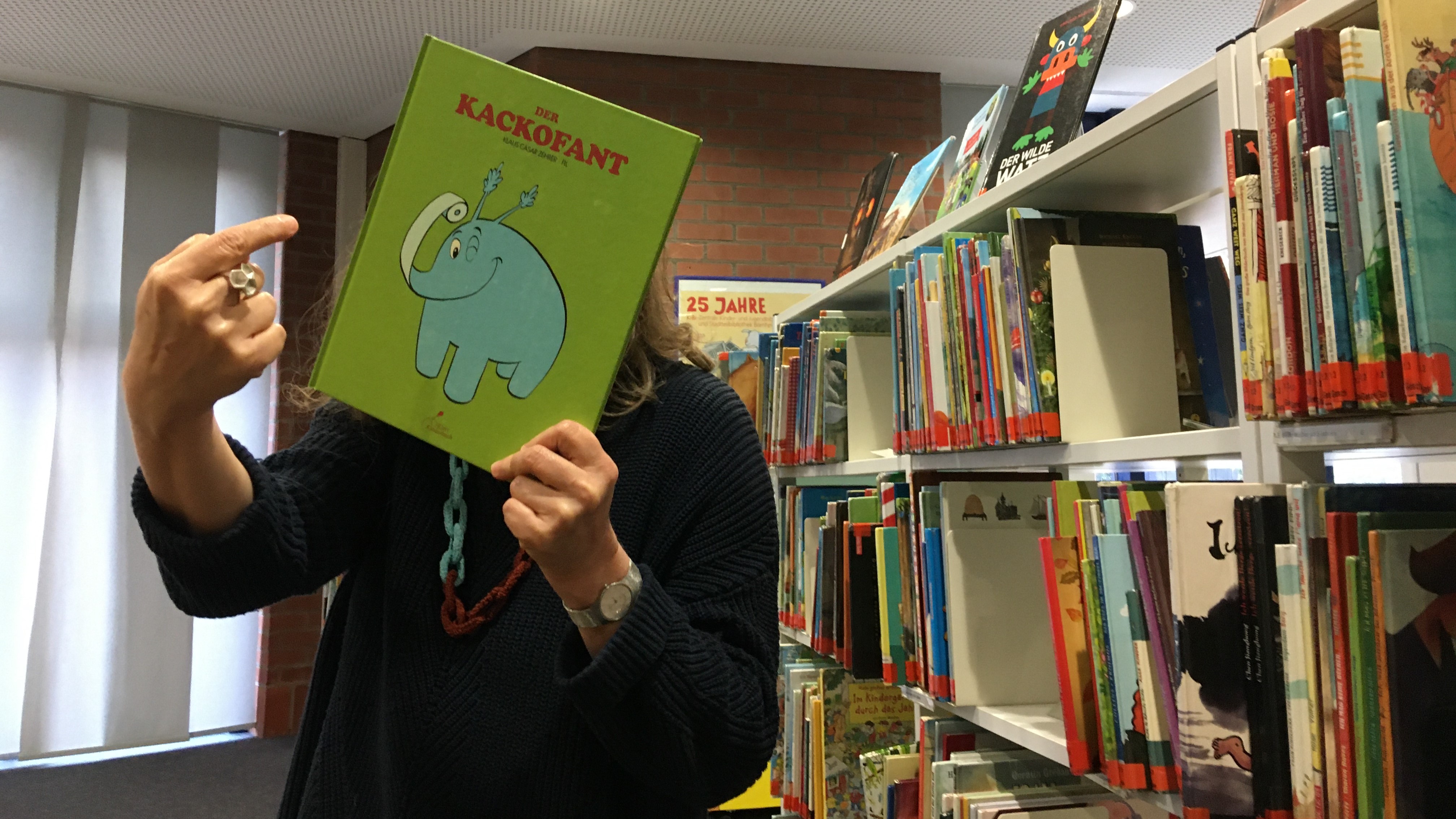 Grünes Buchcover mit blauem Elefanten, hochgehalten von Stadtbüchereimitarbeiterin vor Bücherregal