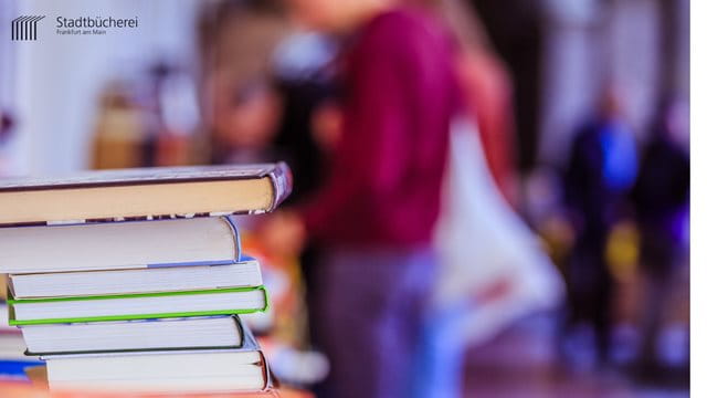 Bücherflohmarkt Bücherflohmarkt. Ein Stapel Bücher steht auf einem Tisch. Im Hintergrund erkennt man eine Frau, auf der Suche nach einem neuen Buch.