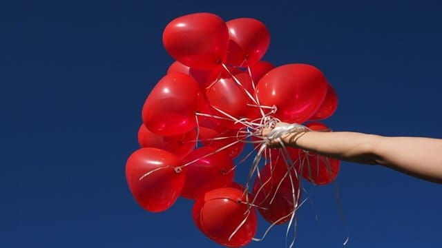 Eine Hand hält viele rote Luftballons in den blauen Himmel.