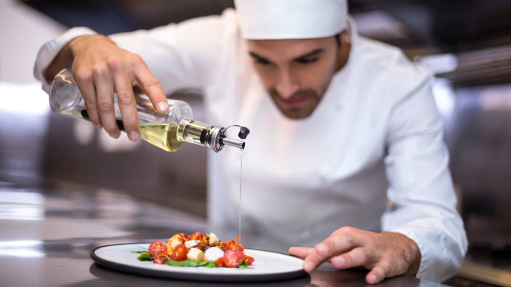 Ein Koch gießt Öl über einen Teller, auf dem Tomaten und Mozarella angerichtet sind.