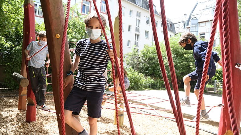 Klettern, rennen, spielen: Mädchen und Jungen mit Mund-Nasen-Bedeckung klettern auf einem Klettergerüst in der Pause.