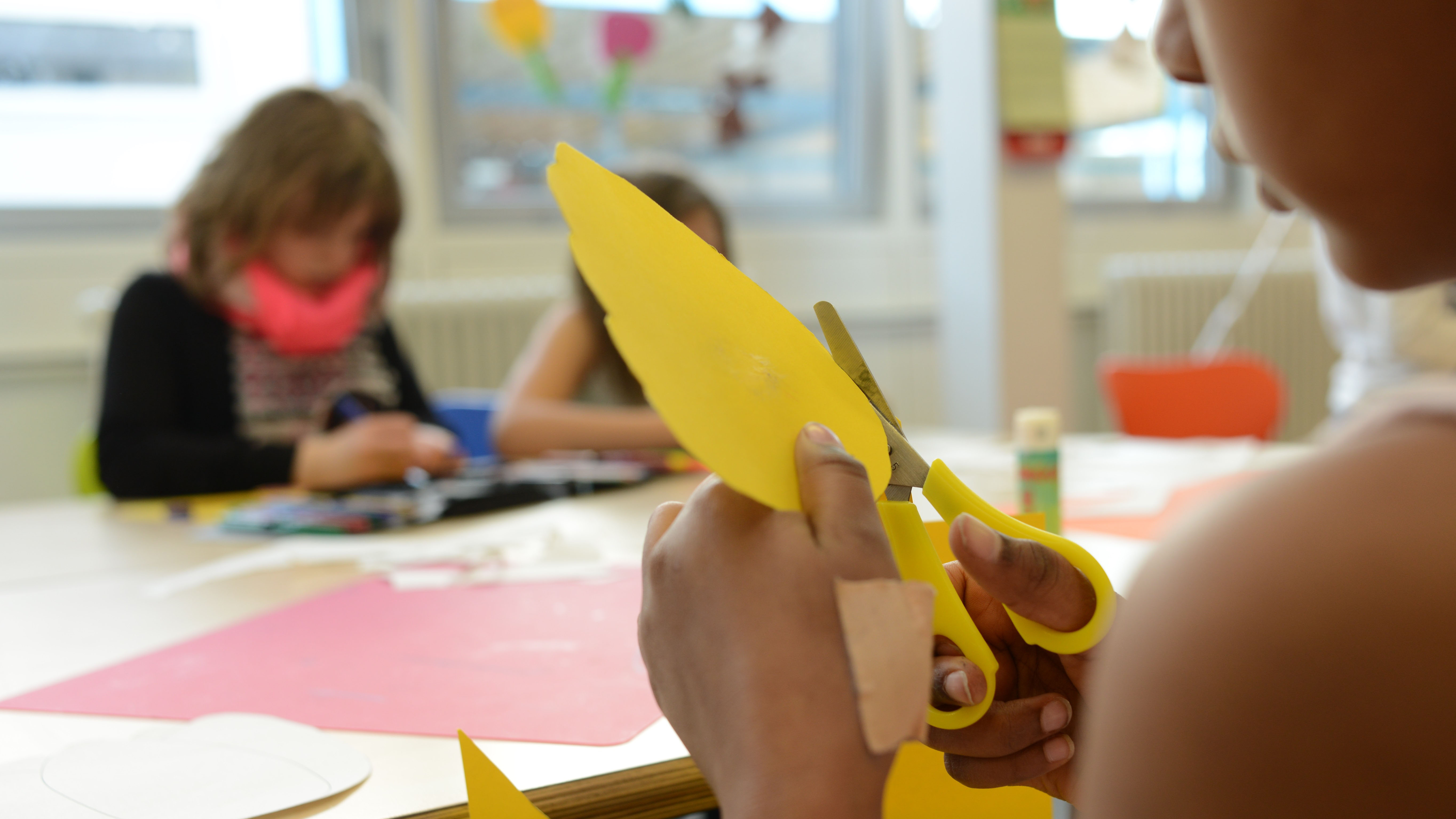 Eine Hand hält eine Schere und schneidet gelbe Pappe. Im Hintergrund Schulkinder.