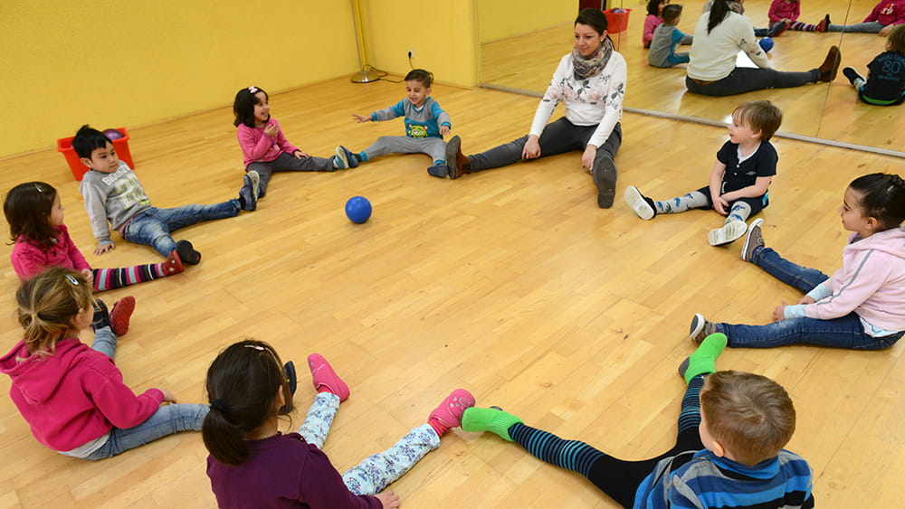 Kinder sitzen mit Betreuungsperson im Kreis, rollen sich den Ball gegenseitig zu.