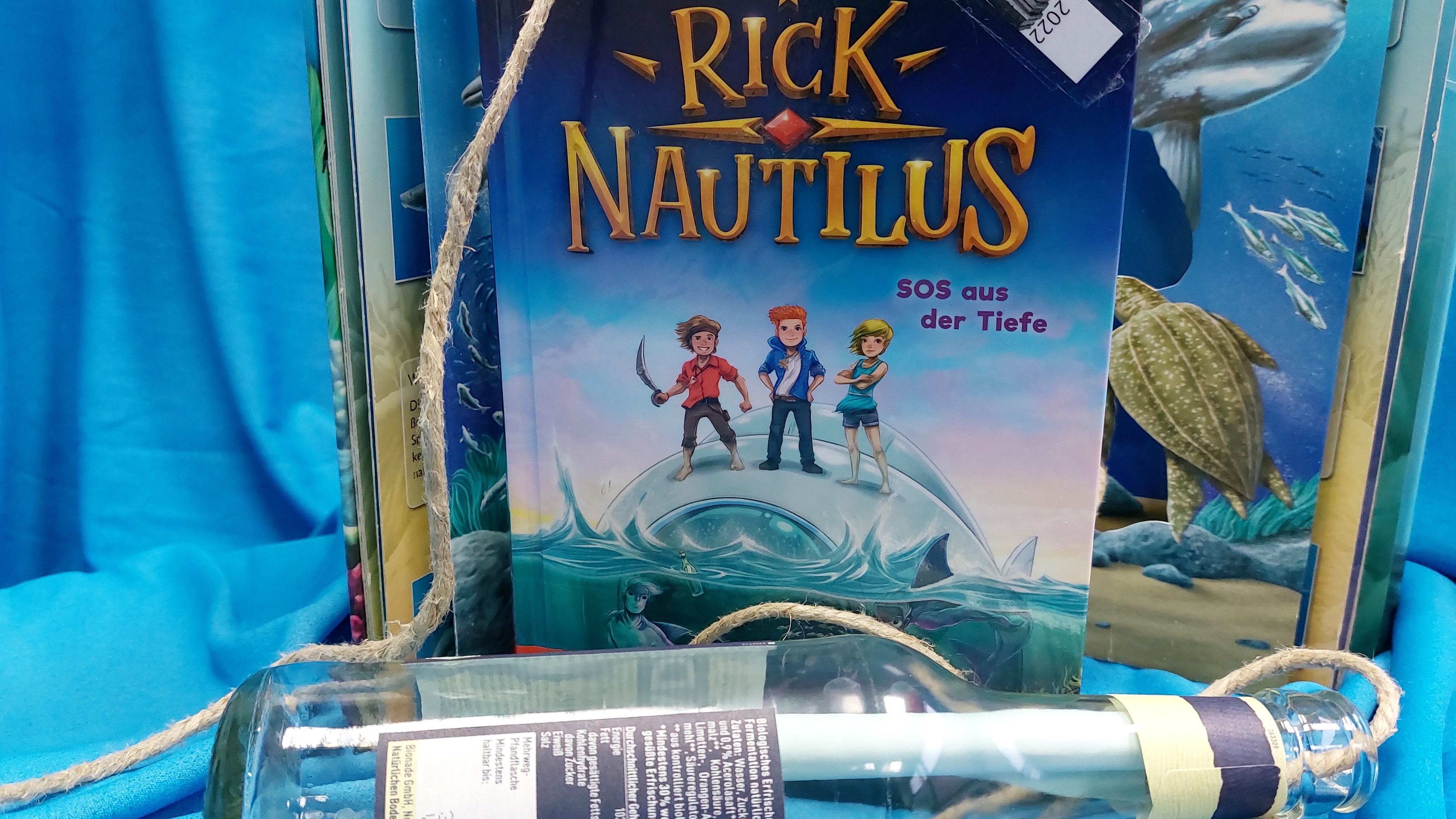 Flaschenpost und Buch 'Rick Nautílus - SOS aus der Tiefe' von Ulf Blanck vor blauem Hintergrund
