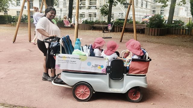 Eine Tagesmutter mit ihren vier Tageskindern, die in einem Bollerwagen sitzen, auf einem Spielplatz.