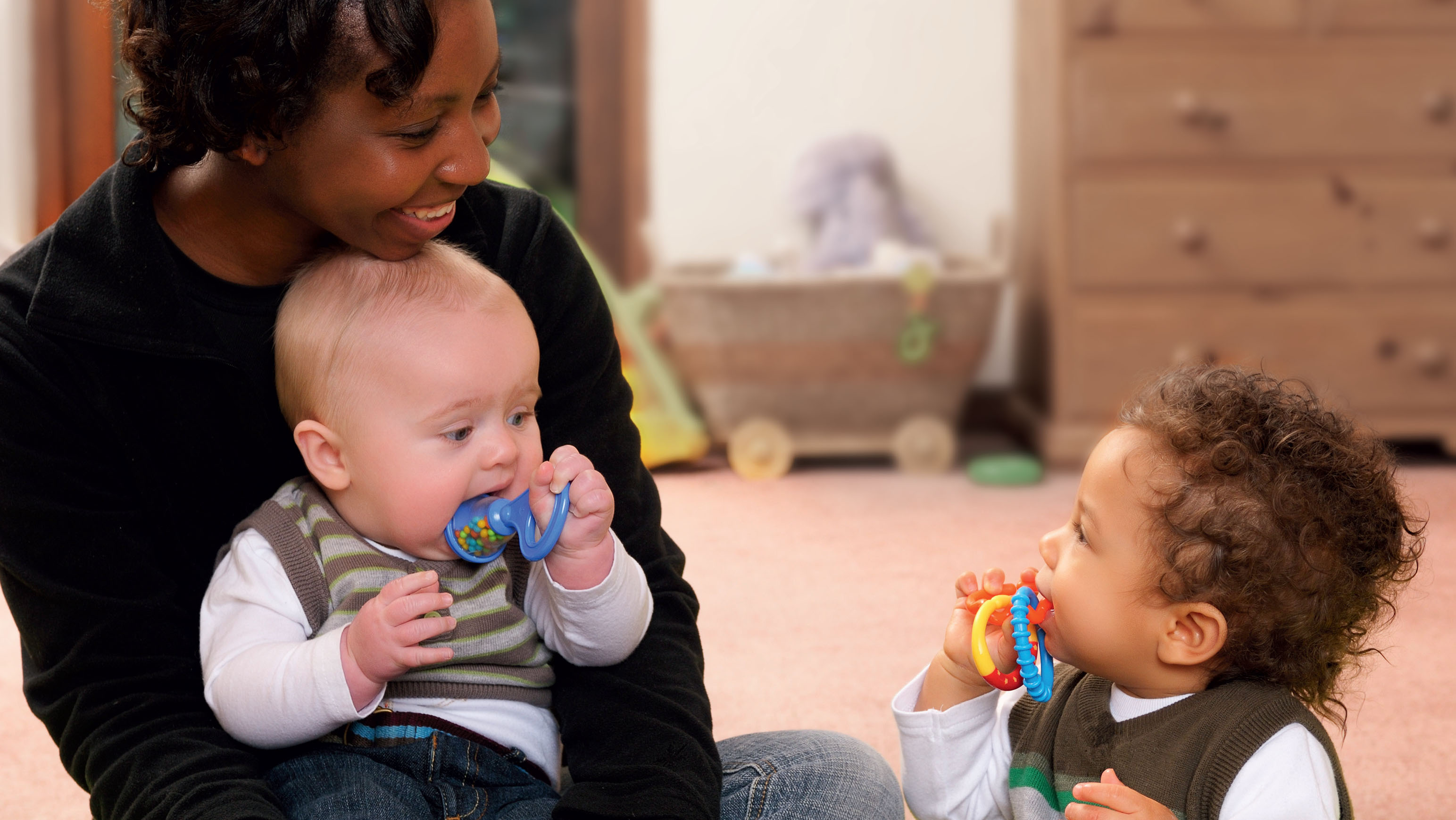 Eine Frau mit einem Baby auf dem Schoß und einem kleinen Kind vor sich sitzen, beide Kinder mit Spielzeug im Mund.