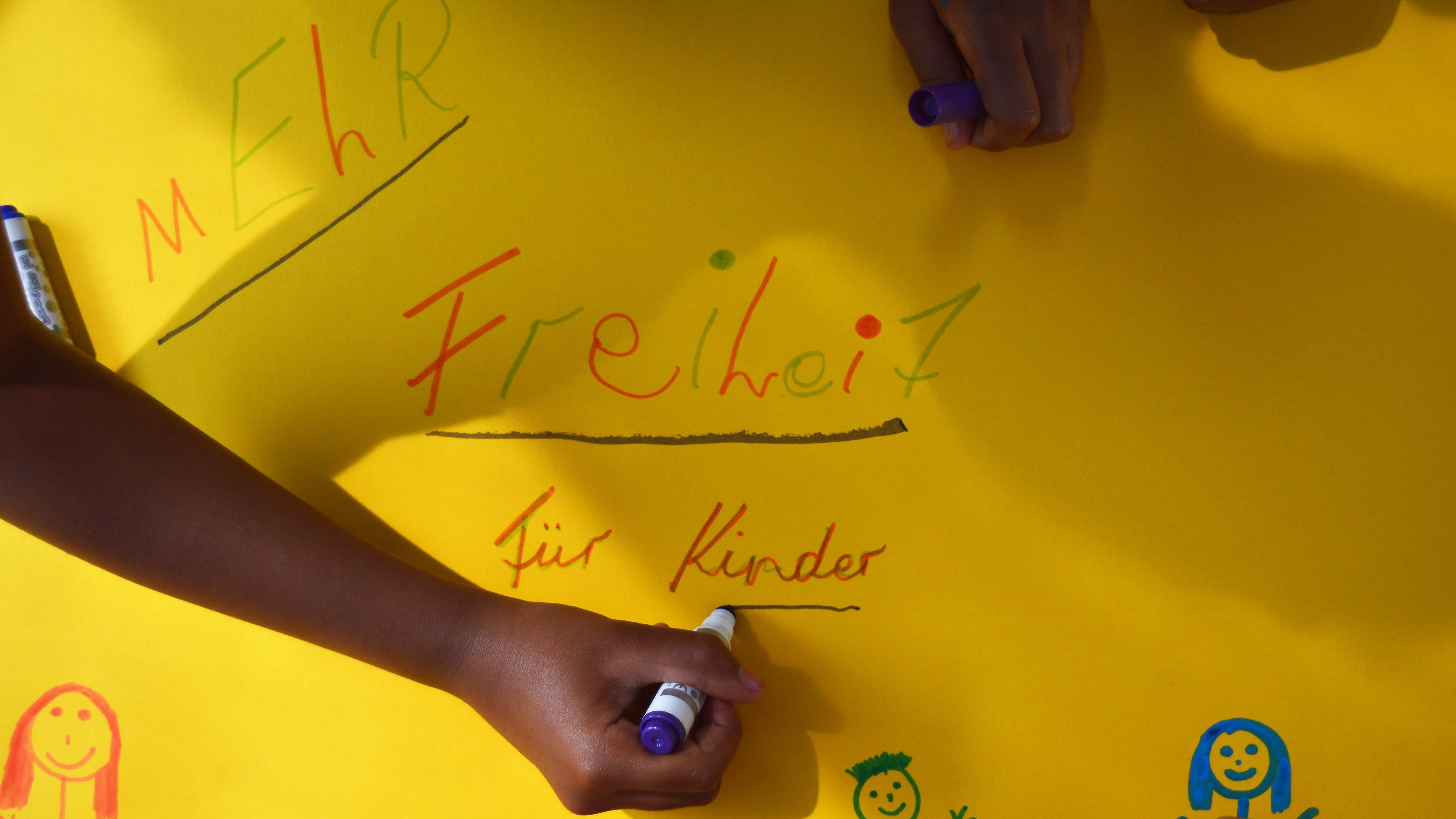 Kinderhände malen und schreiben an einem Plakat mit dem Slogan "Mehr Freiheit für Kinder".